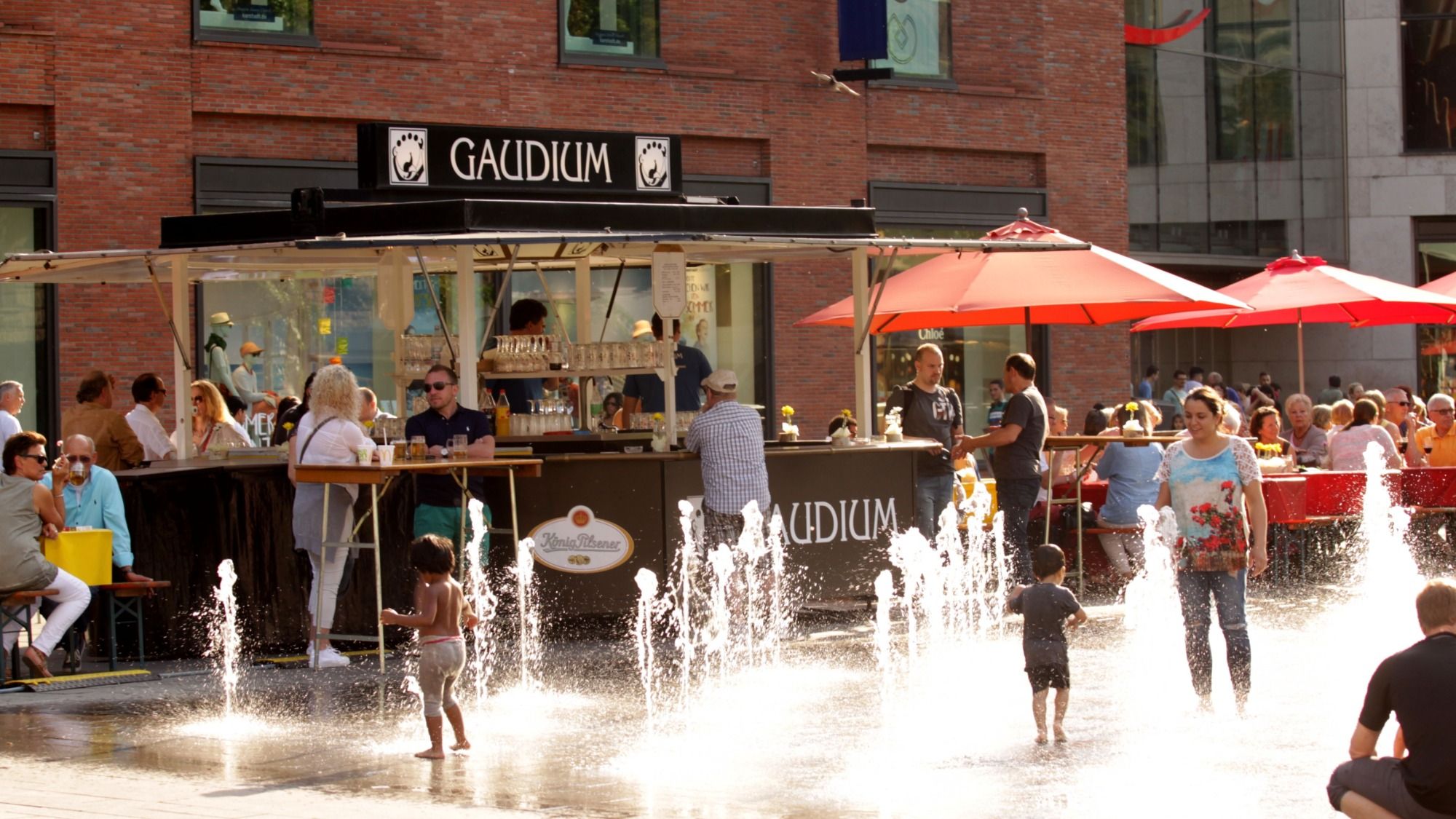 GAUDIUM-Getränkestand mit Logo auf einem sommerlichen Stadtfest – Gäste an Stehtischen und Kinder am Wasserspiel